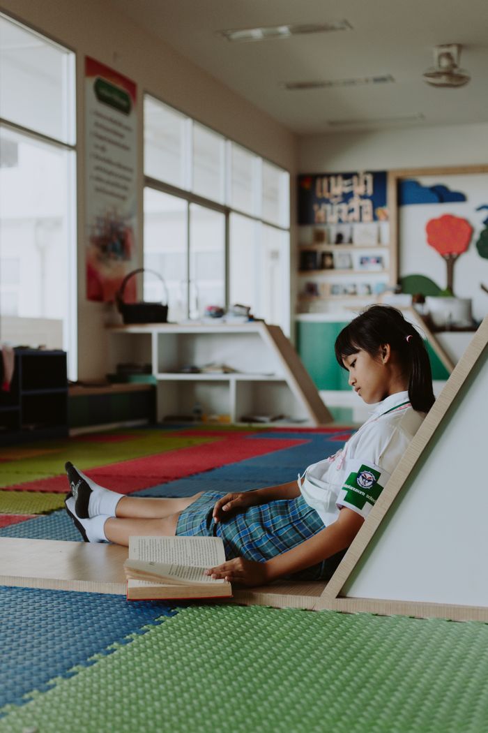A young girl in school uniform reads a book, sitting on the floor in an educational setting.