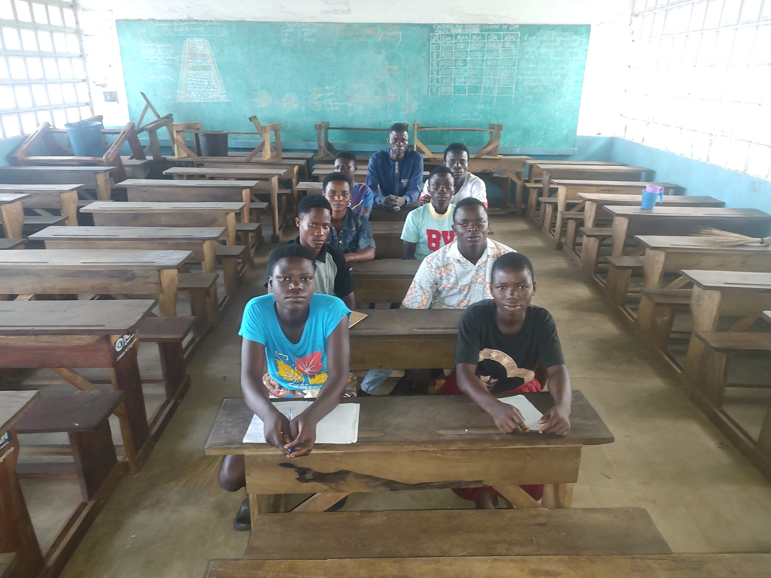 Des élèves et leur enseignant assis à des pupitres en bois dans une salle de classe devant un tableau vert